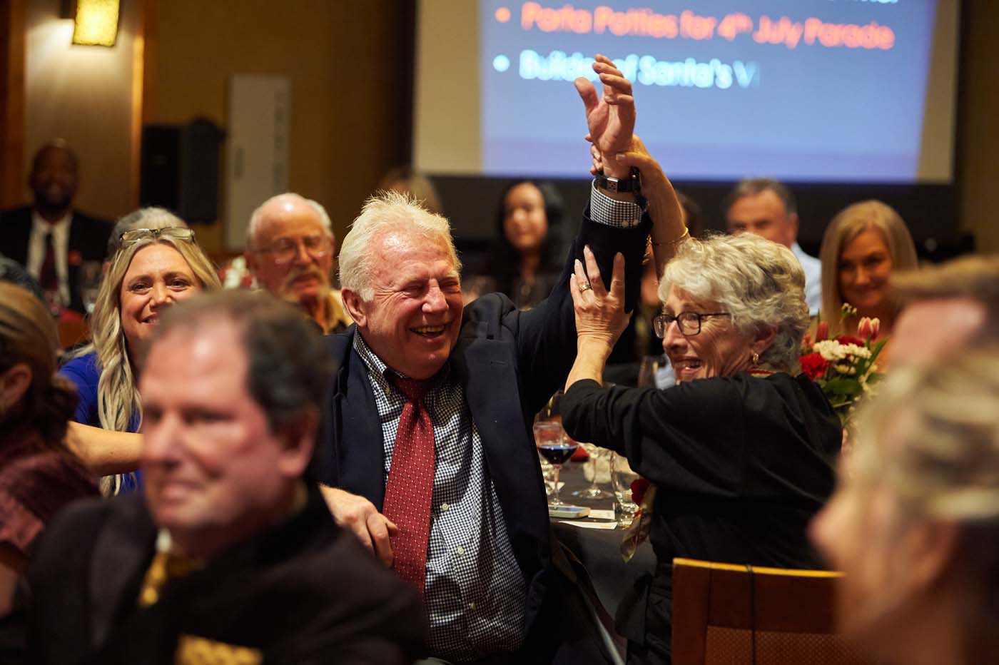 Citizen of the Year Paul Price sitting in a crowded event room, smiling and raising a hand.