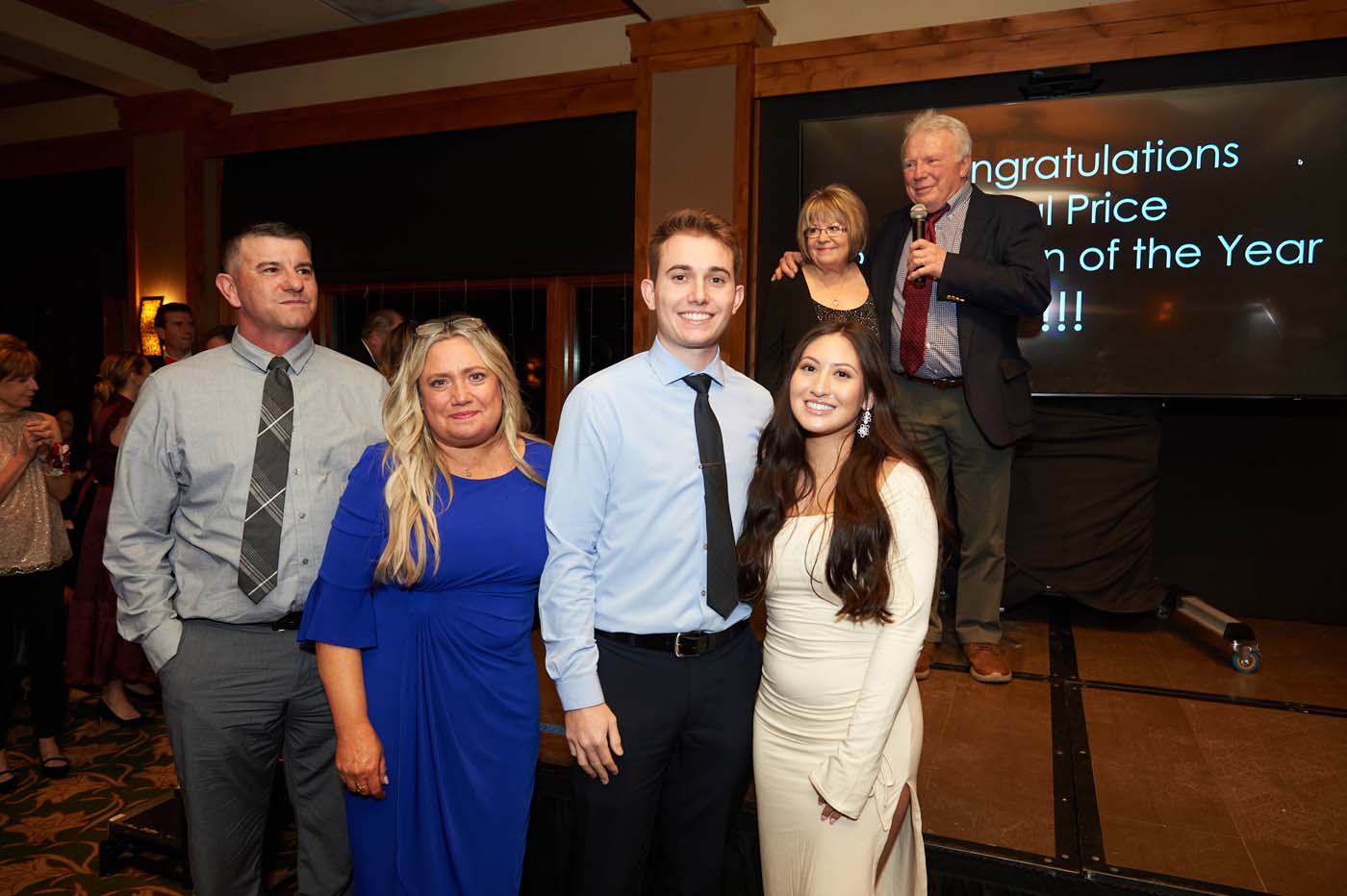 four people standing and smiling, and two people behind them announcing awards on a stage